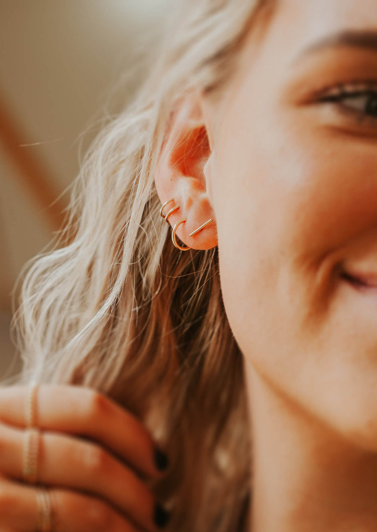 Small gold earrings in Tiny Bar stud style by Hello Adorn styled with twist earrings and gold hoop earrings.