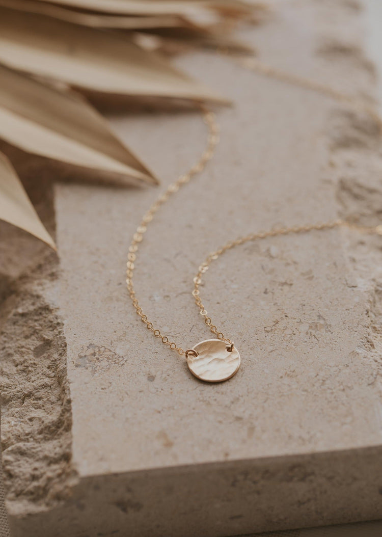 A pendant necklace with a hammered disc attached to a dainty chain necklace to create the Supermoon Necklace by Hello Adorn, a moon necklace with a gold disc necklace.