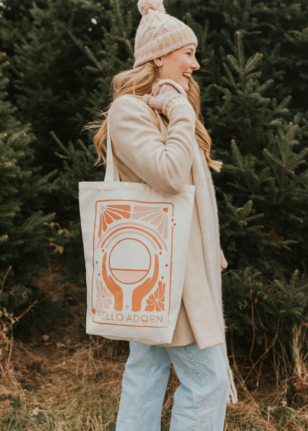 Woman holding a tote bag with a design and text, standing in a forest setting.