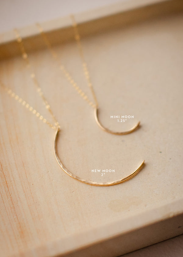 Two crescent moon pendants shown laying flat in two different sizes, one large moon pendant and one small moon pendant attached to a layering chain to create statement necklaces by Hello Adorn.