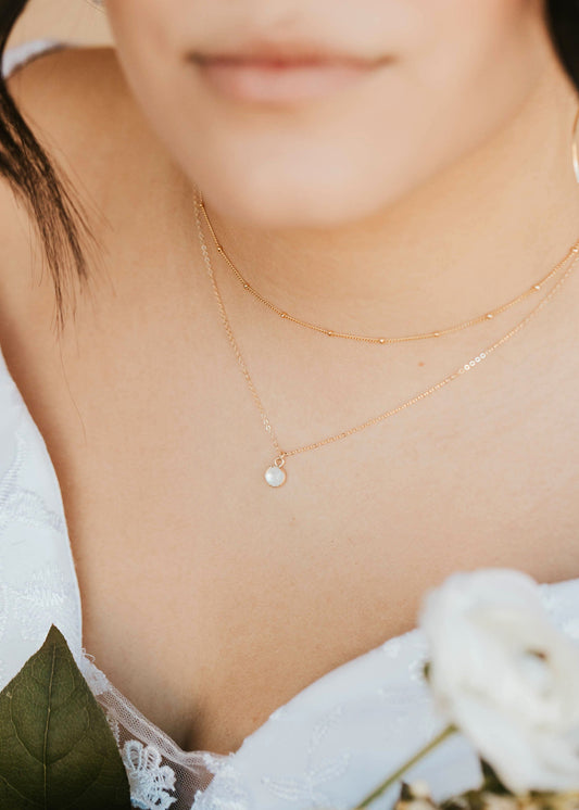 Close-up of a person wearing a delicate gold necklace with a small pendant, holding white flowers.