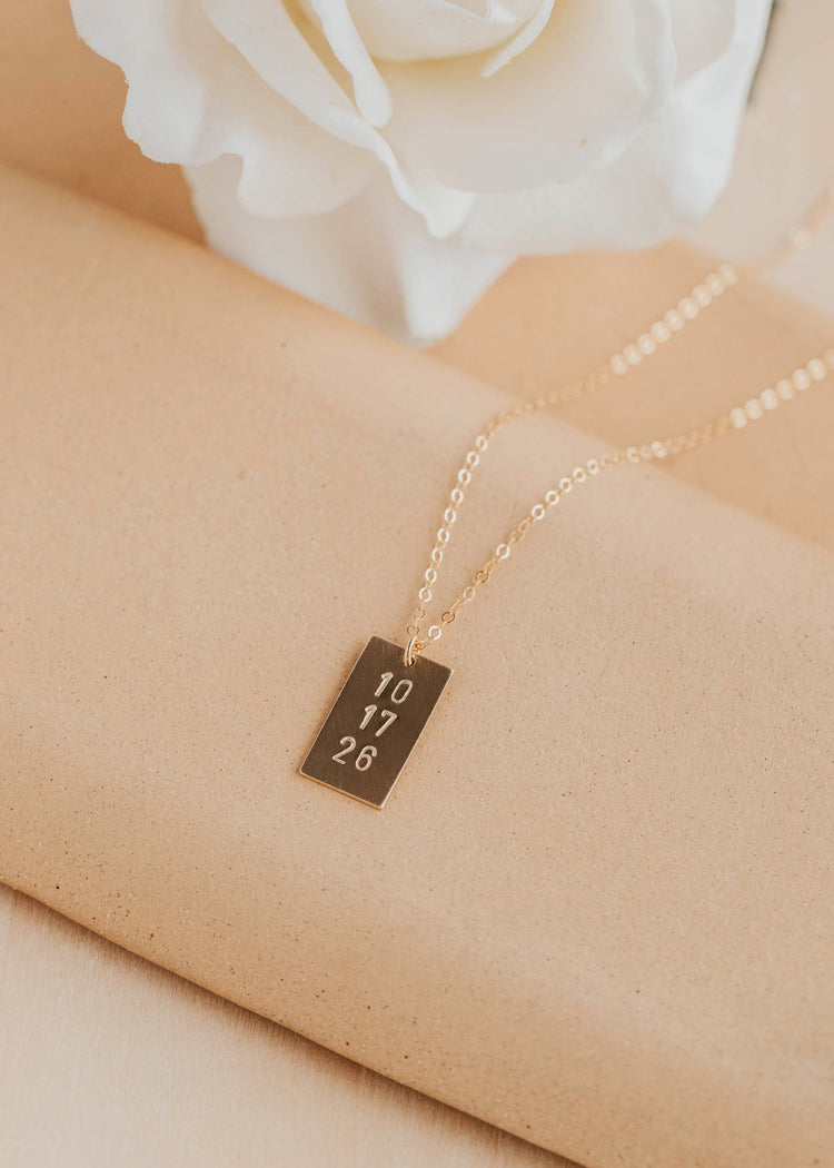 Gold necklace with a bar pendant on a beige surface with a white flower in the background