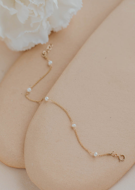 Gold bracelet with pearls on a person's wrist, with a white flower in the background.