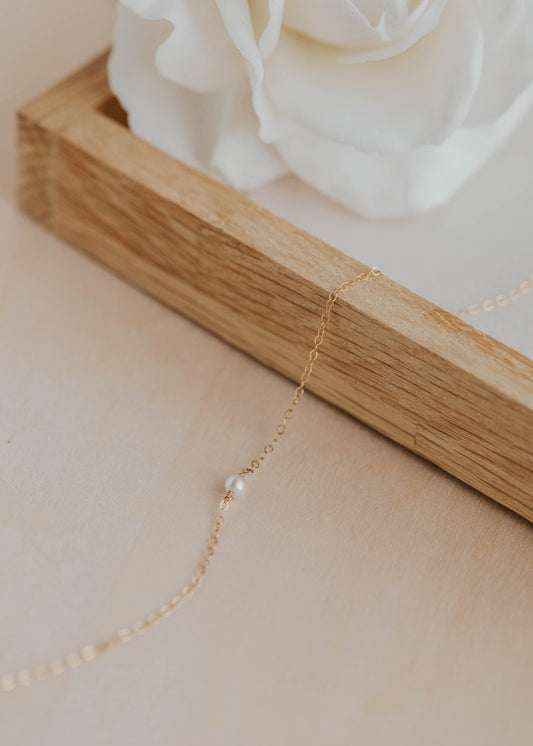 Delicate gold necklace with a pearl pendant on a wooden stand with a white rose in the background.