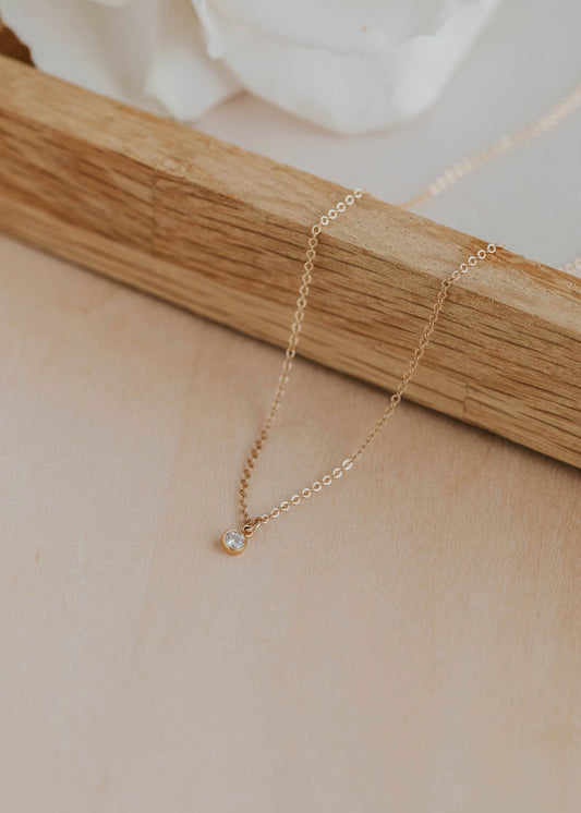Delicate gold necklace with a small clear stone pendant on a wooden stand.
