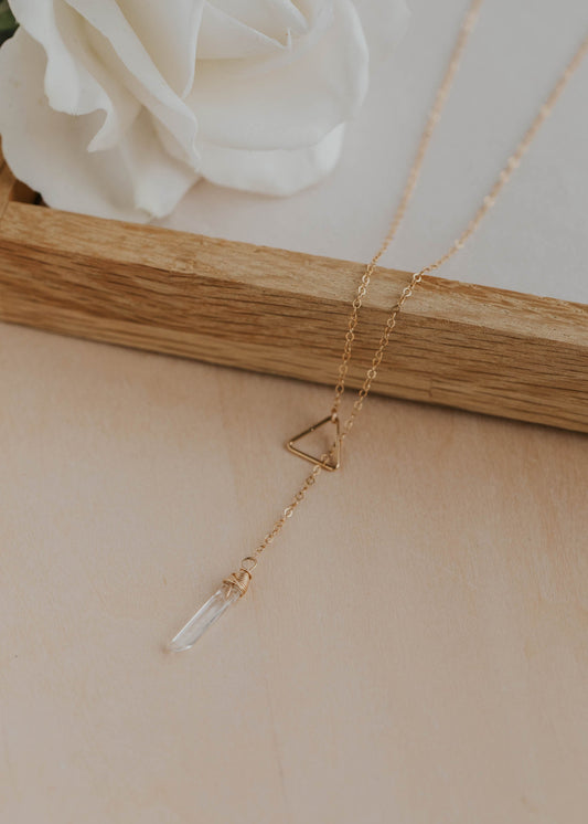 Gold necklace with a triangular pendant on a beige surface with a white flower in the background