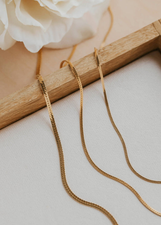 Gold necklaces on a light surface with a wooden frame and white flowers in the background