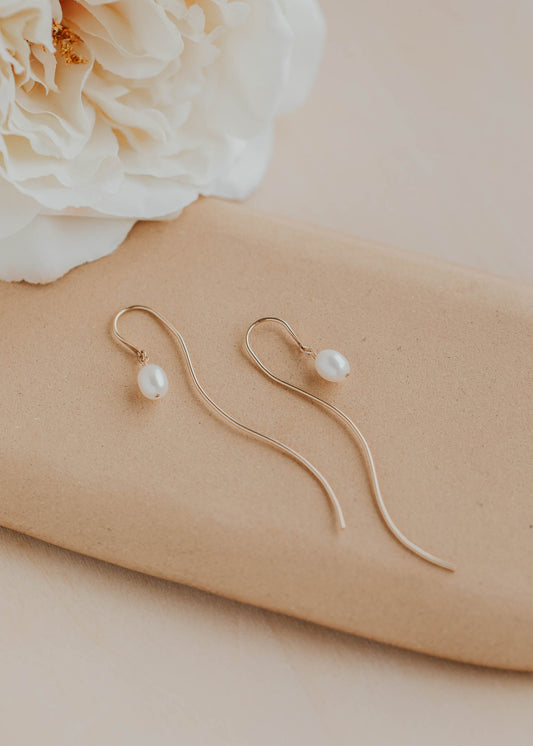 Gold earrings with pearl drops on a beige surface with a white flower.