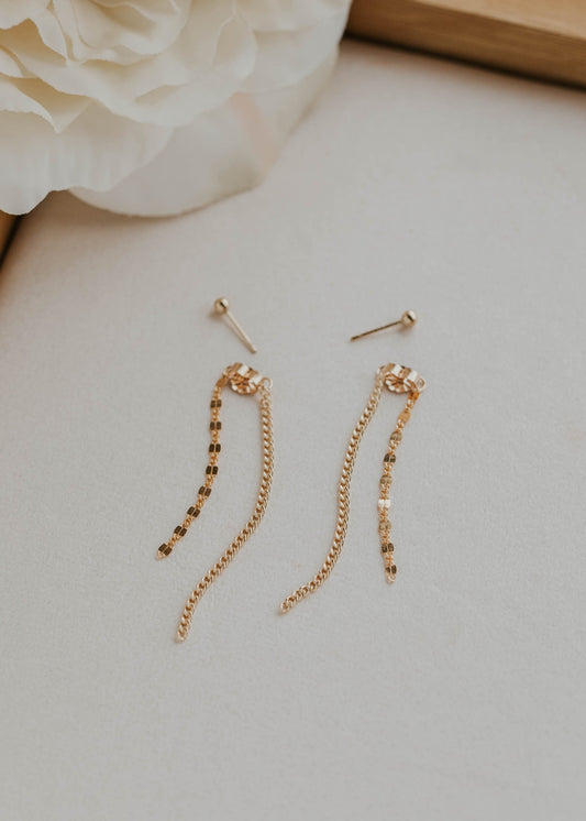 Gold chain earrings on a soft surface with a blurred flower in the background