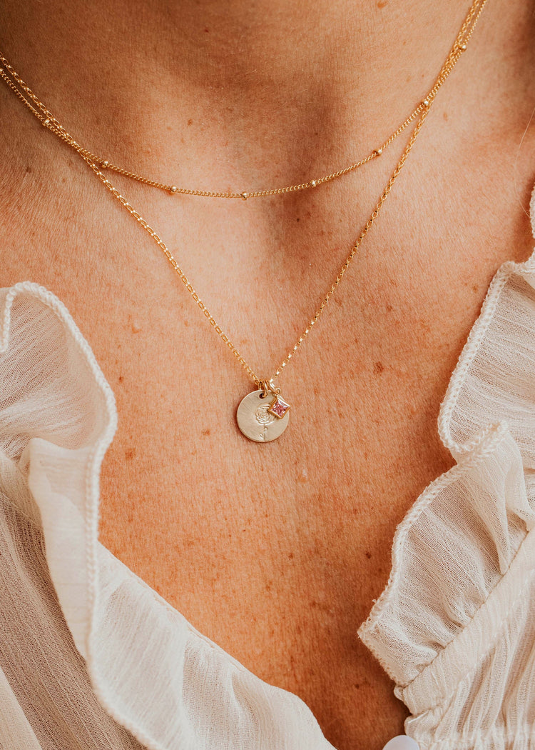 Close-up of a person wearing a Hello Adorn gold necklace with a pendant, with a white blouse in the background.
