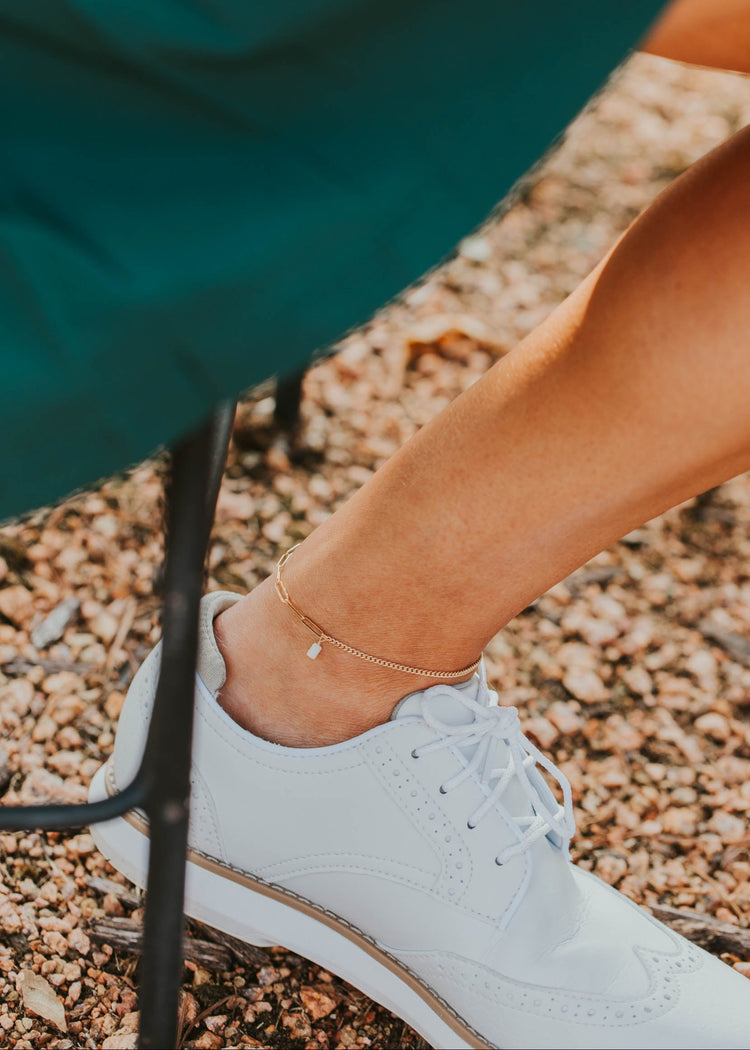 White sneaker on a gravel surface with a green skirt in the background and gold anklet