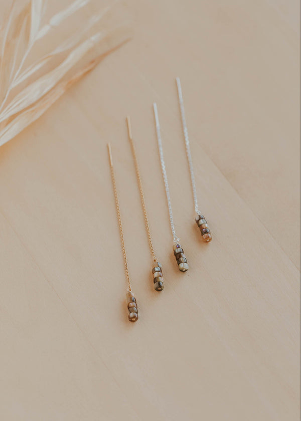 Three delicate earrings on a beige surface with a blurred leaf in the background