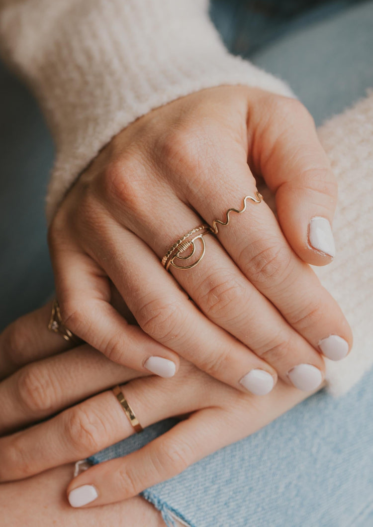 A gold ring stack featuring stacking rings and a statement ring in the Raye Ring style which is a rainbow ring with a double arc and wire wrapped in the center handmade by Hello Adorn.