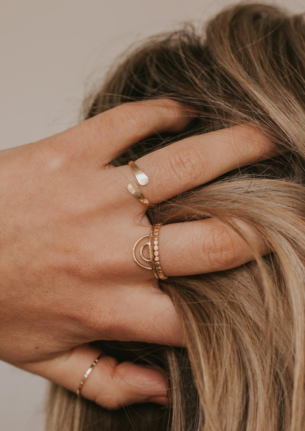 A wrap ring in Sonder design stacked with a rainbow wire wrapping ring in Raye design, hammered beaded band in confetti ring design, and a thumb ring shown in a thin ring band by Hello Adorn all shown in 14k gold fill. 