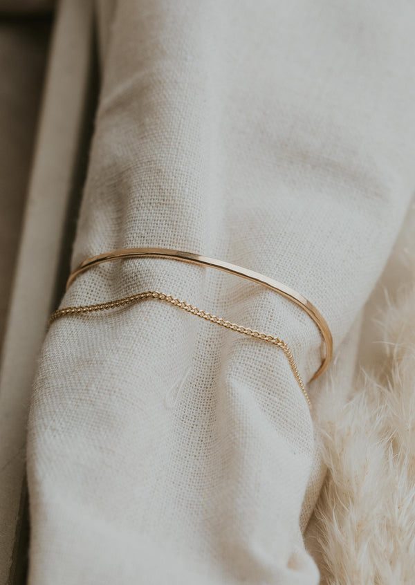 A close up look at a gold cuff bracelet in the Tides Cuff style from Hello Adorn, a simple cuff bracelet with a chain bracelet attached, a staple piece for any bracelet stack.