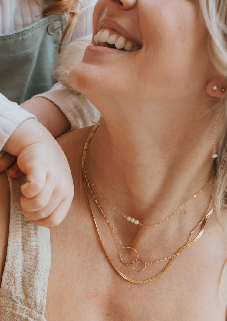 A necklace stack with layering chains and statement necklaces featuring a herringbone chain, interlocking circle necklaces, and a gold pearl necklace from Hello Adorn.