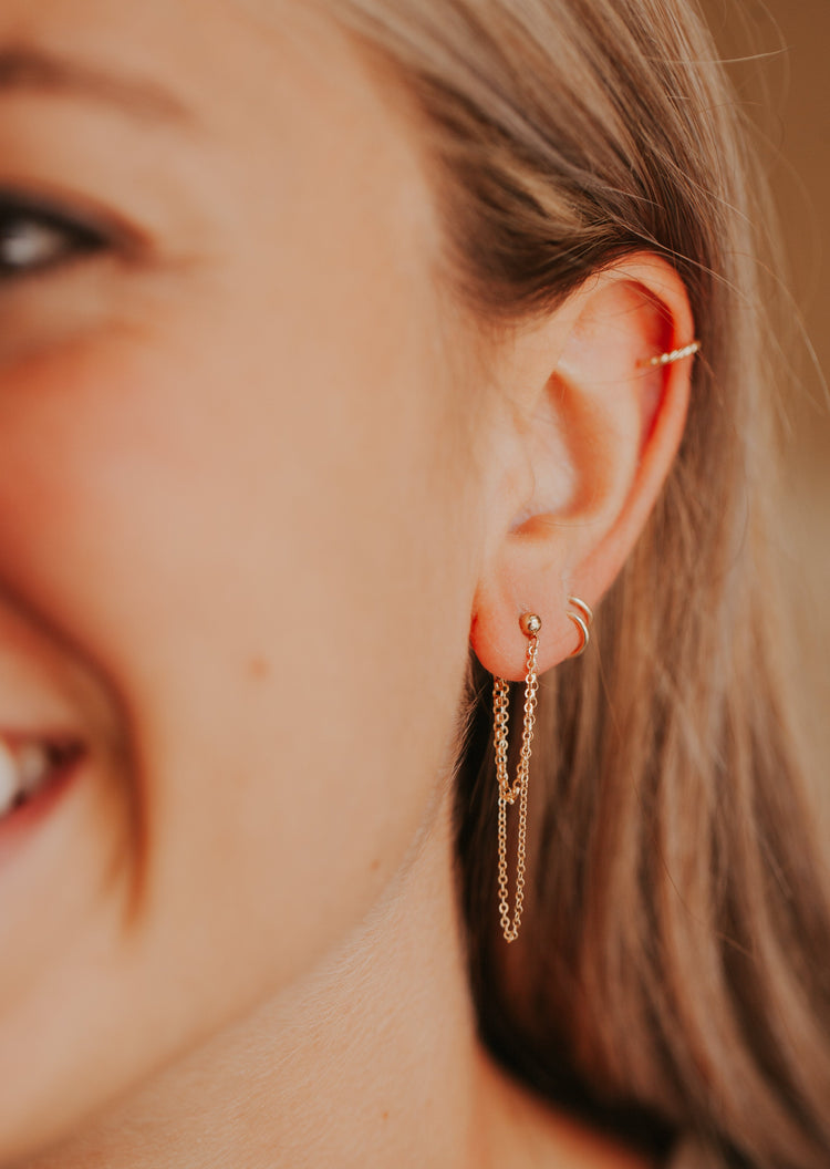 A model wearing Skyla Double Annex earrings created by Hello Adorn, a pair of gold chain earrings combined with dangle earrings and attached to a stud earring paired with twist earrings and an ear cuff.