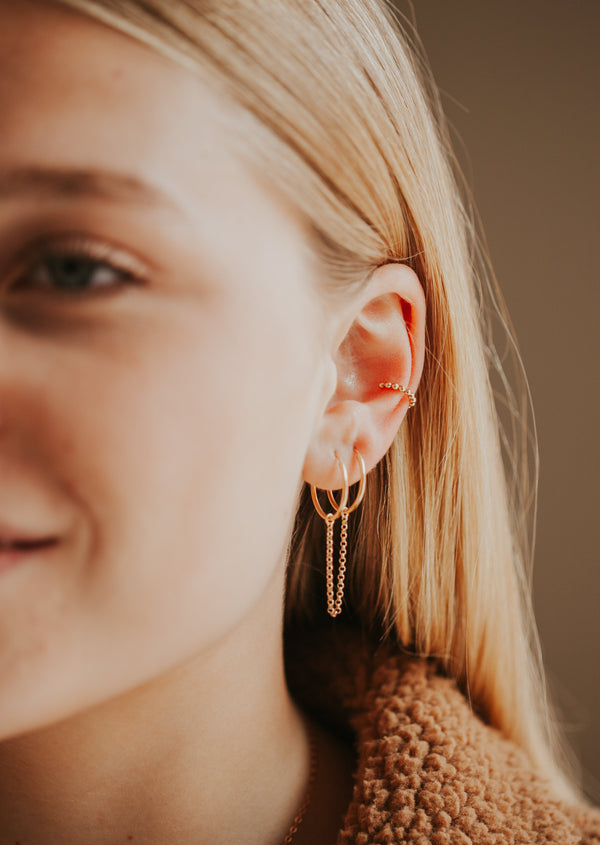 A model wearing a simple earring stack to give you earring stack ideas styled with a gold ear cuff and a dangle chain earring attached to two gold hoop earrings by Hello Adorn.