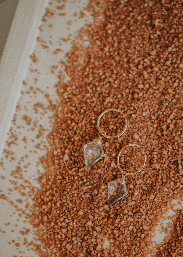 A dagger-shaped, faceted, crystal quartz stone is wire-wrapped and attached to a 14kt Gold Fill endless hoop earring flat laying in clay-colored sand.