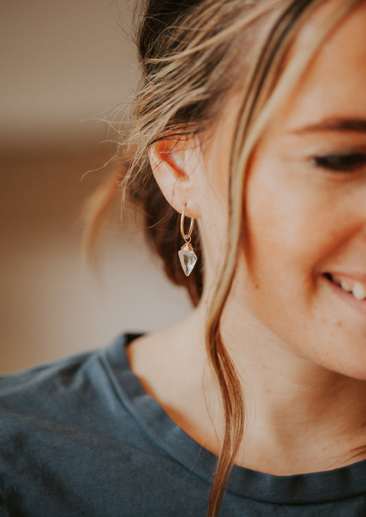 A dagger-shaped, faceted, crystal quartz stone is wire-wrapped and attached to a 14kt Gold Fill endless hoop earring is modeled on a woman. 