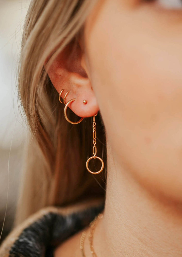 An earring stack in 14k gold fill featuring Hello Adorn's Monday Backdrop which is a dangle earring attached to a stud earring with a circle attached at the end of a chain styled with a hoop earring and a twist earring.