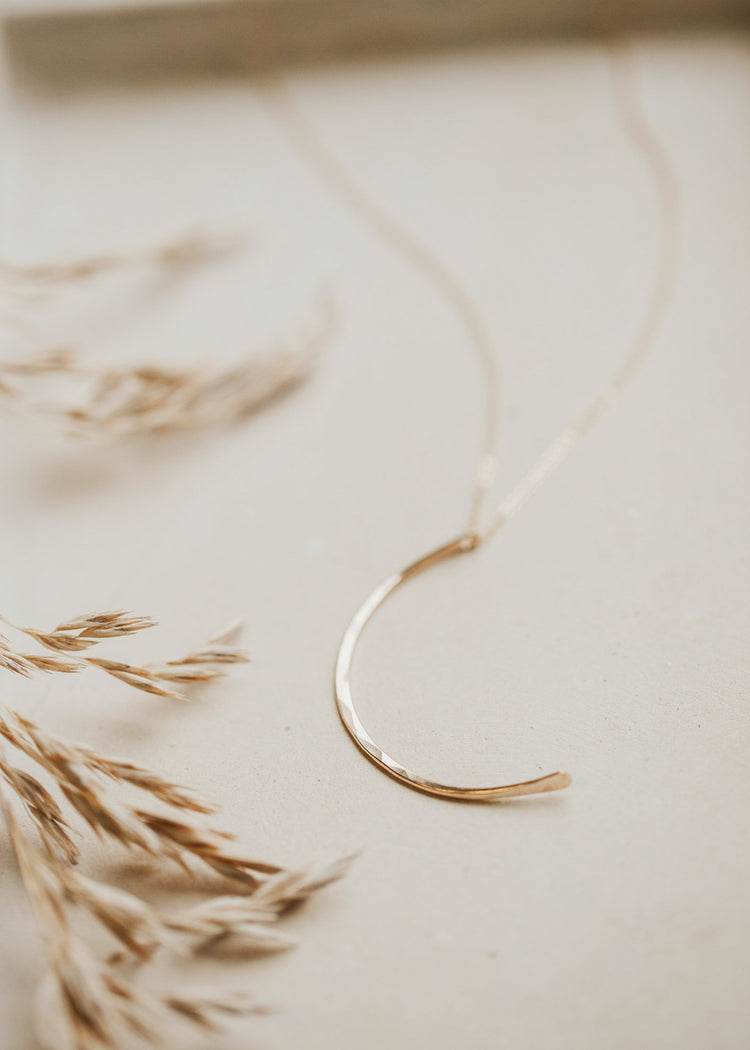 A moon necklace in the shape of a crescent moon attached to a chain necklace using a hammered gold pendant by Hello Adorn in 14k gold fill.