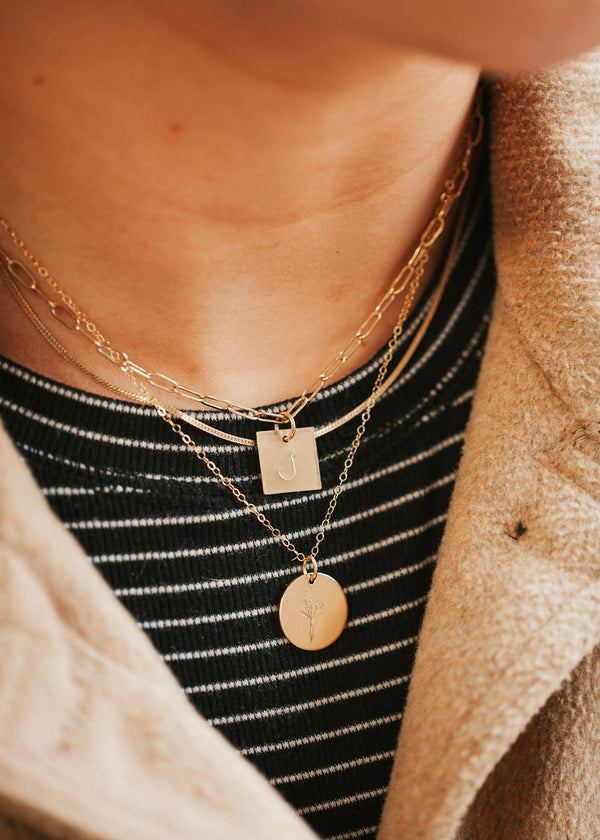 A necklace layering idea with a hand-stamped initial necklace attached to a linked necklace, a herra chain, and a necklace with flower pendant created by Hello Adorn. 