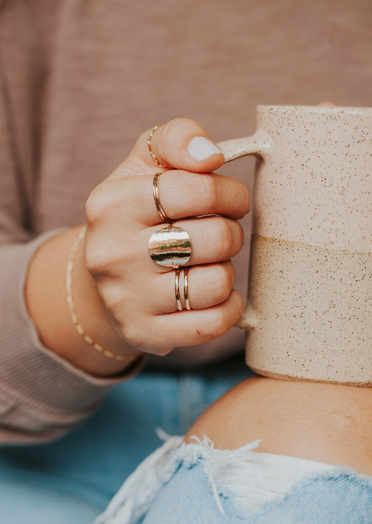 A gold ring stack shown on a model wearing a wrap ring to look like 2 rings, a Supermoon Ring as the statement ring to look like a moon ring, a thin band, a thin hammered ring, and a braided ring styled by Hello Adorn shown in 14k gold fill.