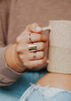 A gold ring stack shown on a model wearing a wrap ring to look like 2 rings, a Supermoon Ring as the statement ring to look like a moon ring, a thin band, a thin hammered ring, and a braided ring styled by Hello Adorn shown in 14k gold fill.