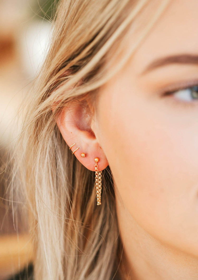 A model showing off her earring stack styled with a gold ball earring, twist earrings, and an earring with a chain shown in 14k gold fill by Hello Adorn.