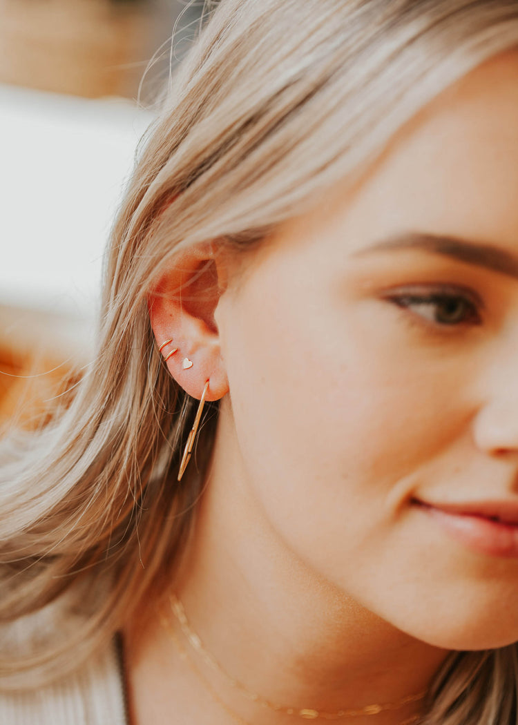 Young woman wearing small heart studs in 14kt Gold Fill layered with open heart slide earrings and tiny twist earrings.